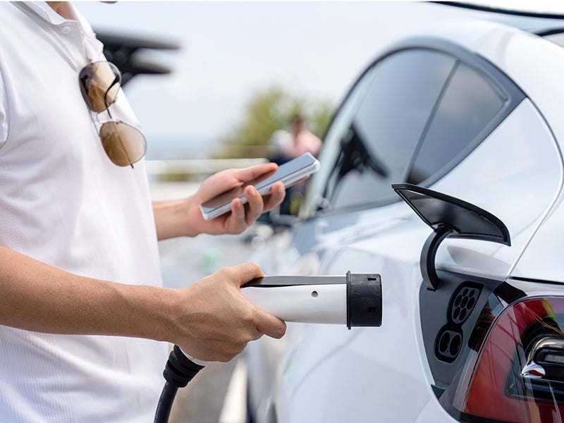 A man in a white polo shirt holds a phone and an EV charging plug near a white electric car.