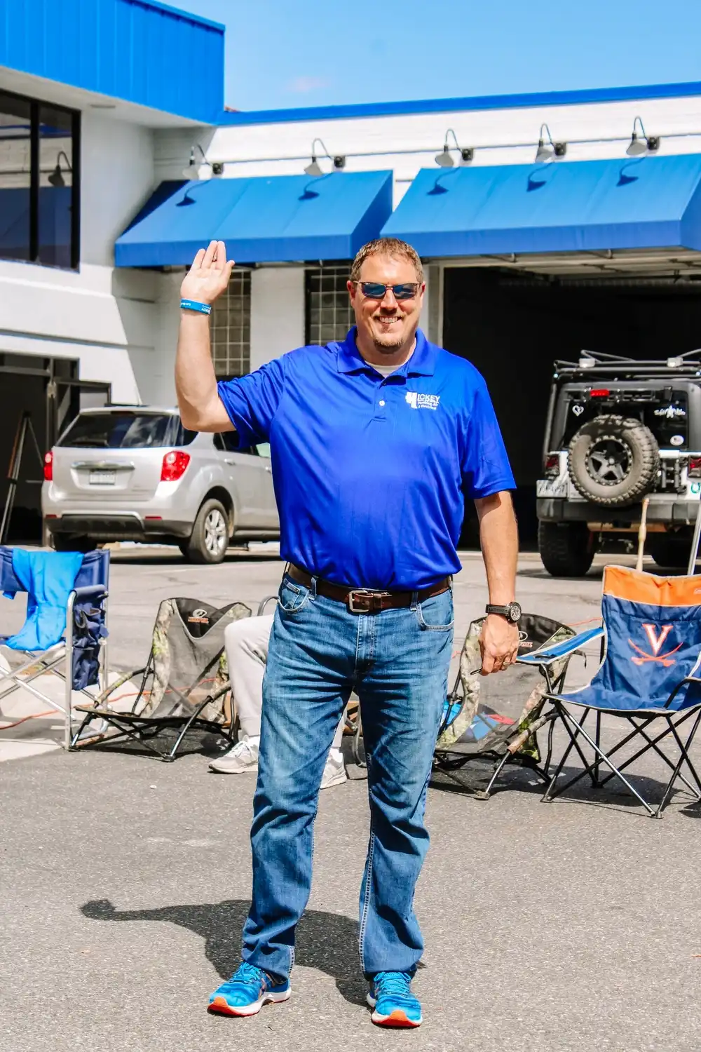 A smiling man in sunglasses, blue polo, and jeans waves while standing in a parking lot.
