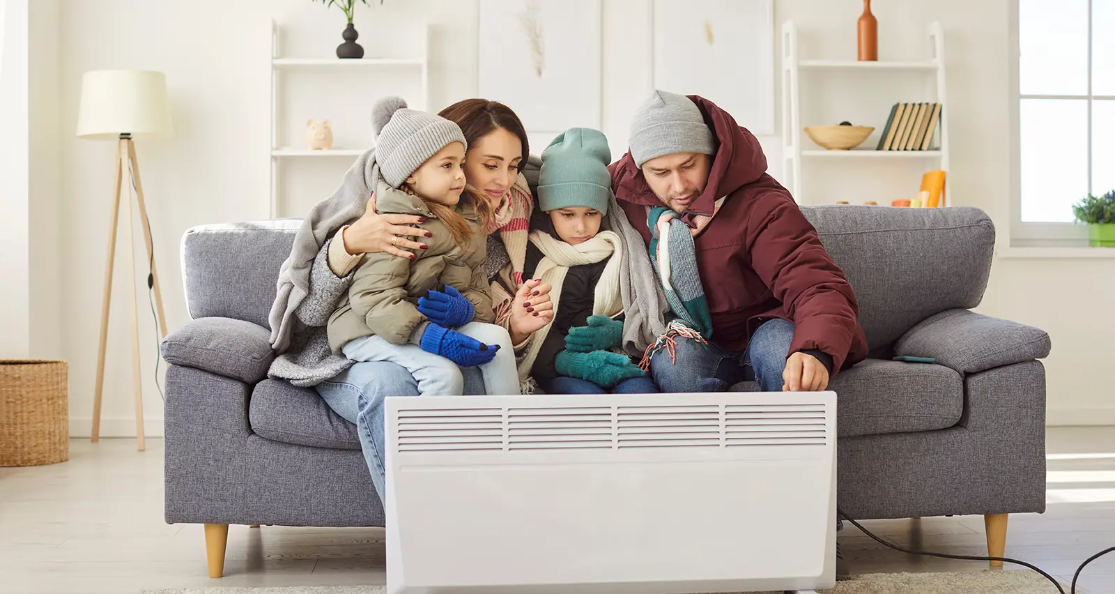 A family of four, bundled in winter clothes, gathers around a space heater on a couch.