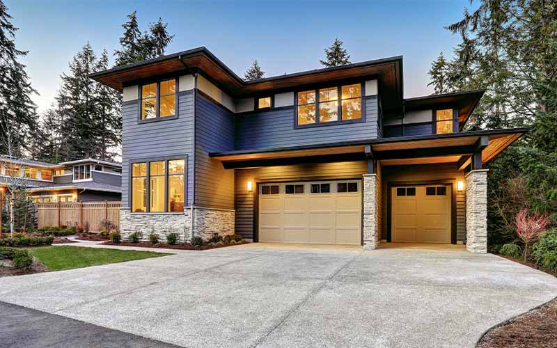 A modern gray and brown house with two garages, a concrete driveway, and green landscaping under a clear sky.