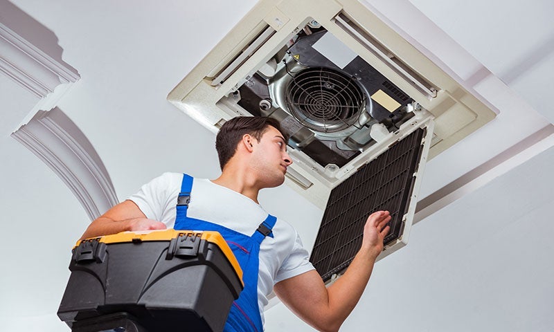 An HVAC technician in overalls inspects an open ceiling air conditioner unit, holding a black and yellow toolbox.