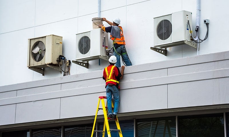 Two workers maintain outdoor air conditioners on a building. One pours from a bucket, while the other stands on a ladder.
