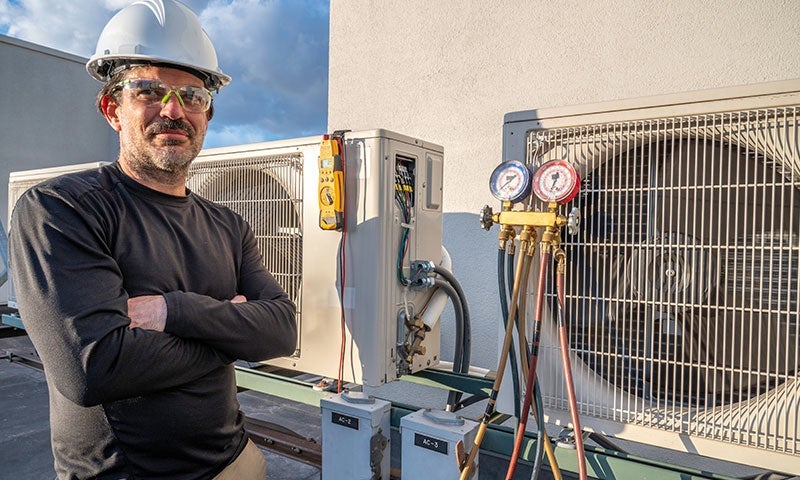 A man in a hard hat and safety glasses stands with crossed arms next to outdoor HVAC units with gauges attached.