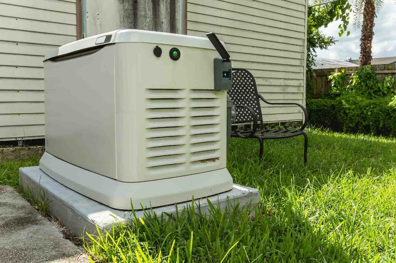 A large, off-white generator sits outside a house, next to a black metal chair and green grass.