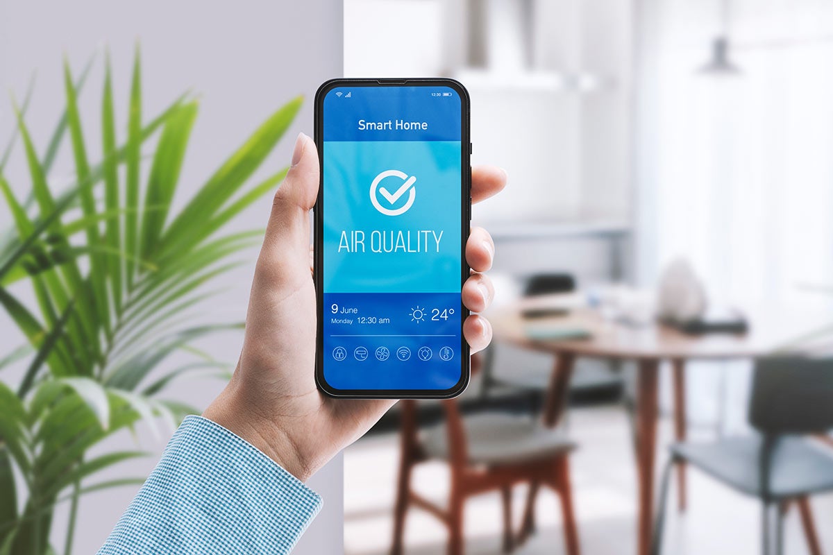 A hand holds a smartphone displaying a smart home app with 'AIR QUALITY' and a checkmark, 24°C, Monday, June 9, 12:30 AM over a blue background. The background is a blurred room with a plant and a table.