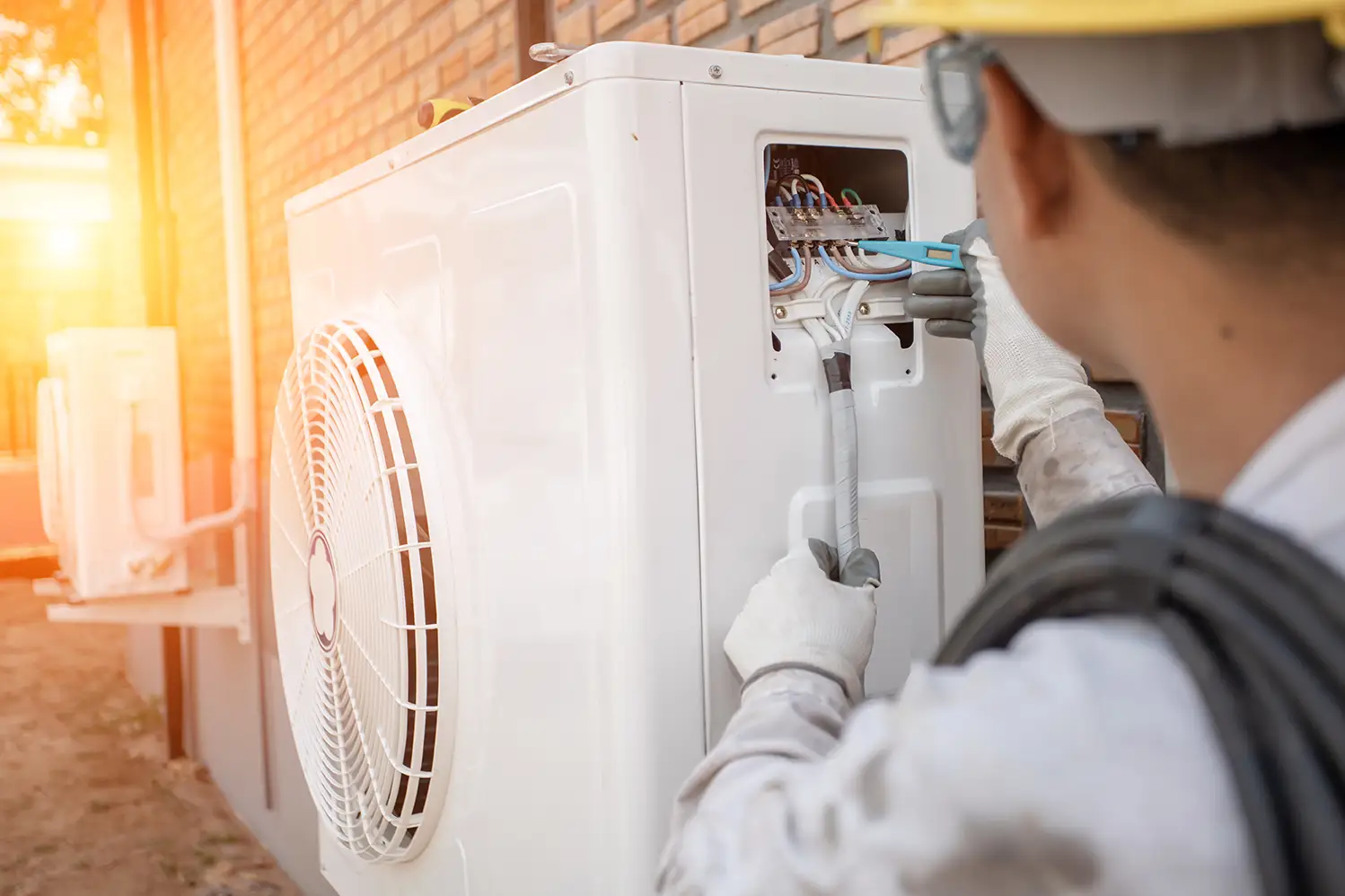 A technician in a hard hat and gloves works on the exposed wiring of an outdoor air conditioner unit.