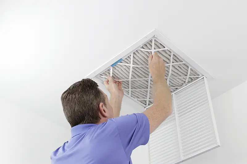 A man in a blue shirt changing a dirty air filter in a white ceiling vent.