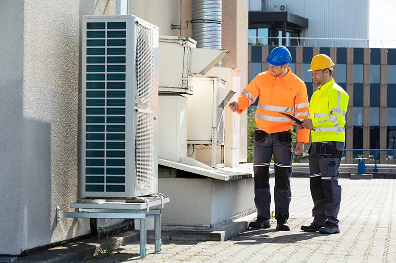 Two technicians in hard hats and reflective jackets inspect outdoor HVAC units on a rooftop.