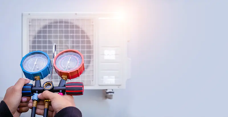 Hands using red and blue gauges to check an air conditioner unit on a white wall.