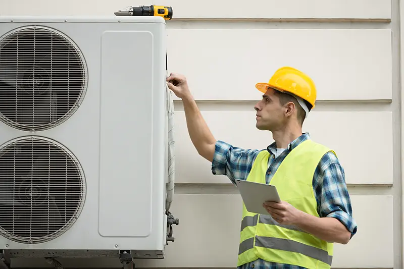 A technician in a hard hat and safety vest maintains an outdoor air conditioning unit.