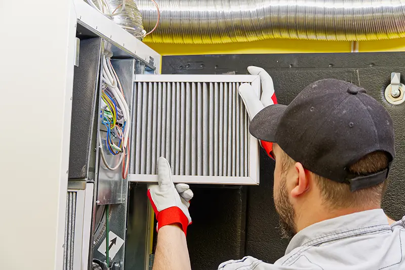 A person wearing a black cap and gloves inserts a new HVAC filter into a unit with exposed wiring.