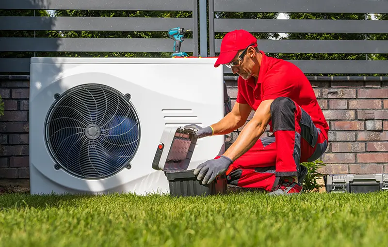Technician in red uniform and safety glasses, checking a white outdoor heat pump unit next to a brick wall.