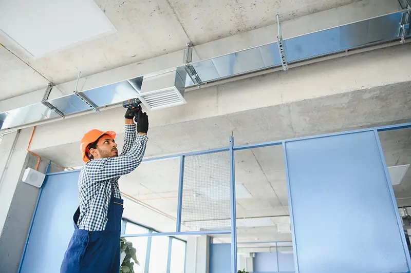 A worker in an orange hard hat and blue overalls uses a drill to install a metallic air duct on a concrete ceiling.