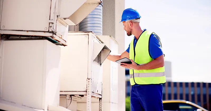 A man in a hard hat, safety vest, and uniform checks air vents on an outdoor unit, holding a tablet.
