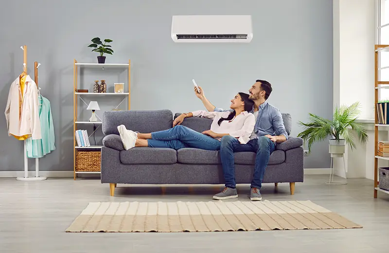 A happy couple sits on a gray sofa, looking up at an air conditioner while holding a remote control.