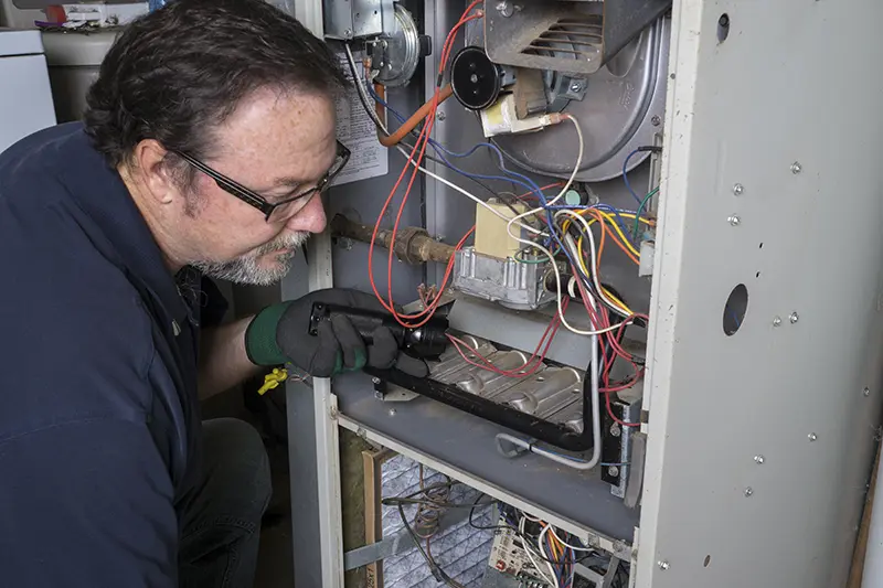A gloved technician with glasses and a beard inspects the exposed wiring and components inside an open furnace.