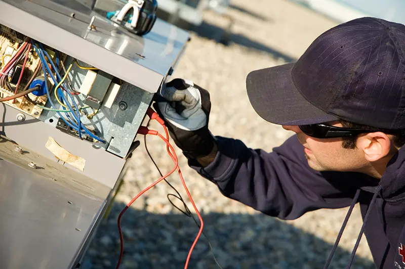 A technician in a baseball cap and sunglasses checks wiring inside an HVAC unit with an electrical tester.
