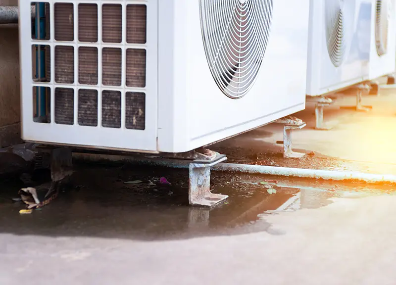Multiple outdoor air conditioning units on rusty stands, dripping water into puddles on the ground.