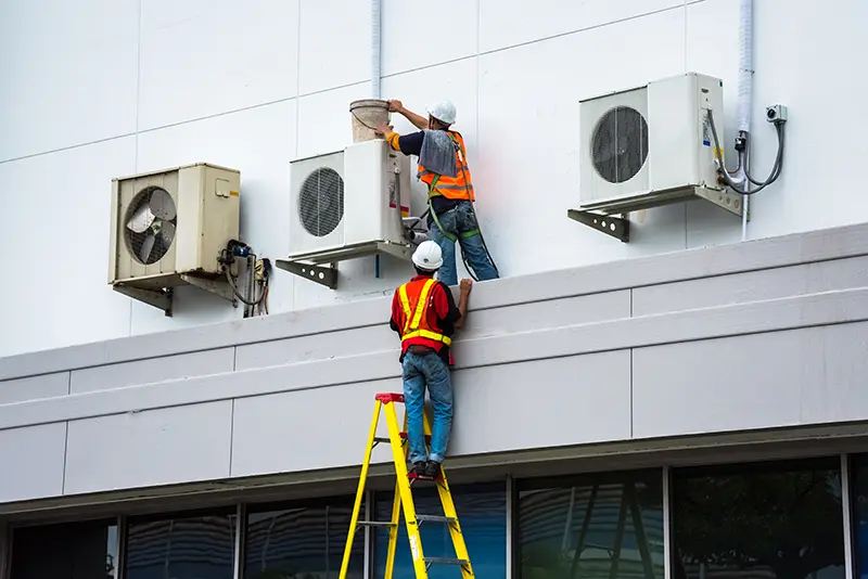 Two HVAC technicians, one on a ladder, maintain air conditioning units on a building wall.