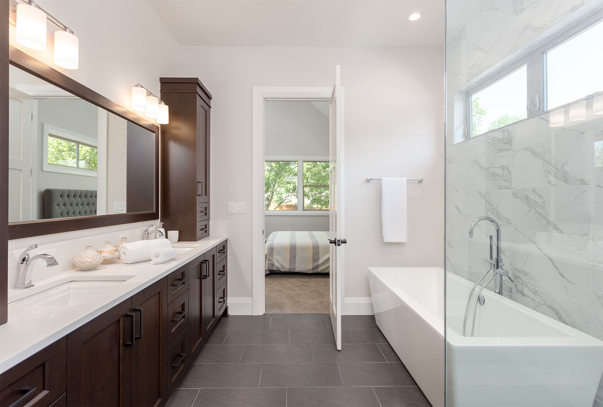 A modern bathroom with dark wood cabinets, dual sinks, a large mirror, a white freestanding tub, and a bedroom visible through an open door.