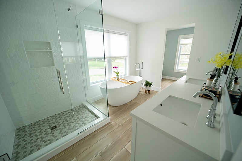 Bright, modern bathroom with a glass shower, freestanding tub by a window, and double vanity with white countertops.