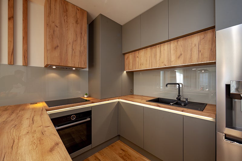 A modern kitchen with wood grain and gray cabinets, light wood countertops, a black cooktop, oven, and sink.