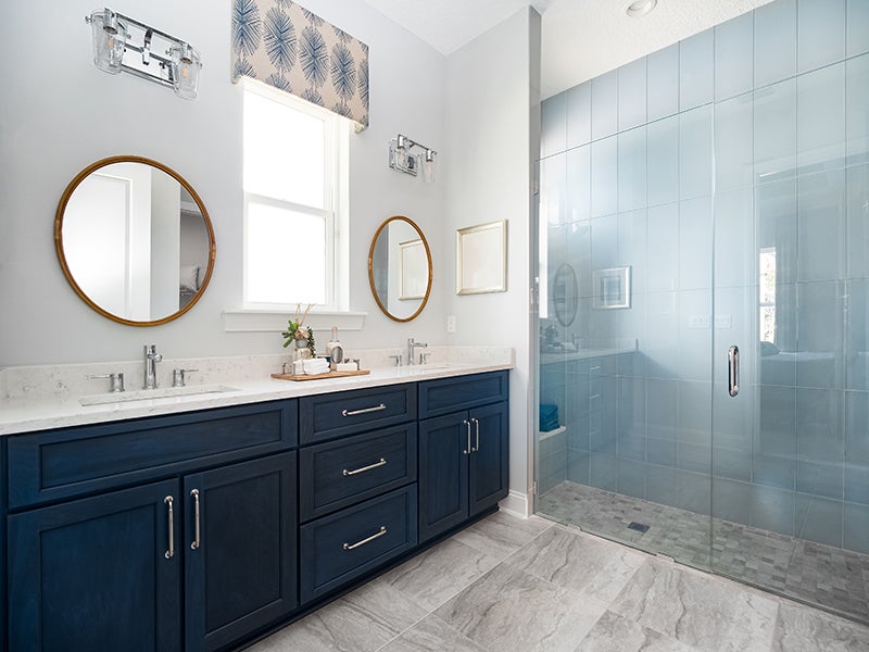 Modern bathroom with double navy blue vanity, gold framed mirrors, light gray walls, and a large glass shower.
