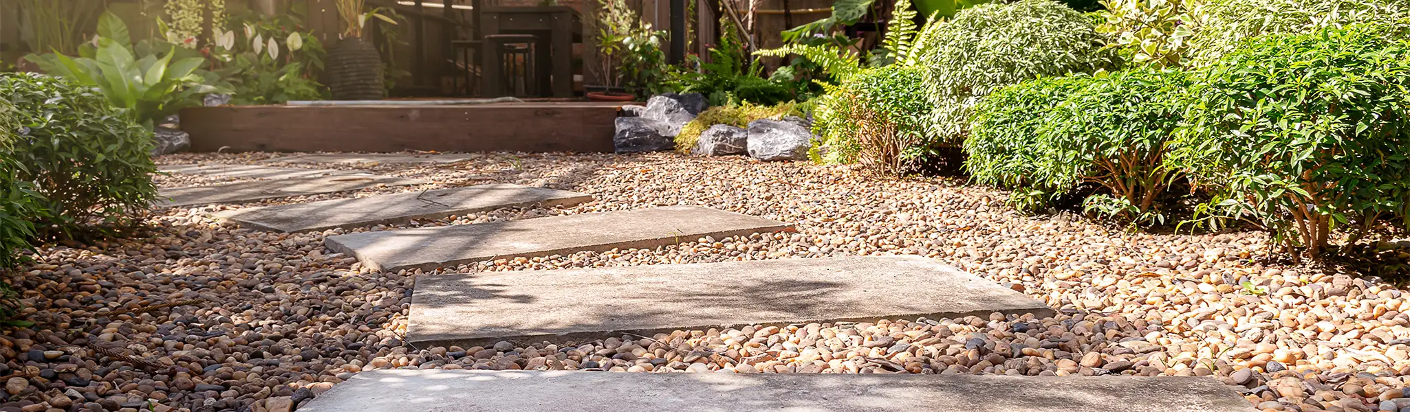Concrete stepping stones across a pebble path through a garden with green plants and shrubs.