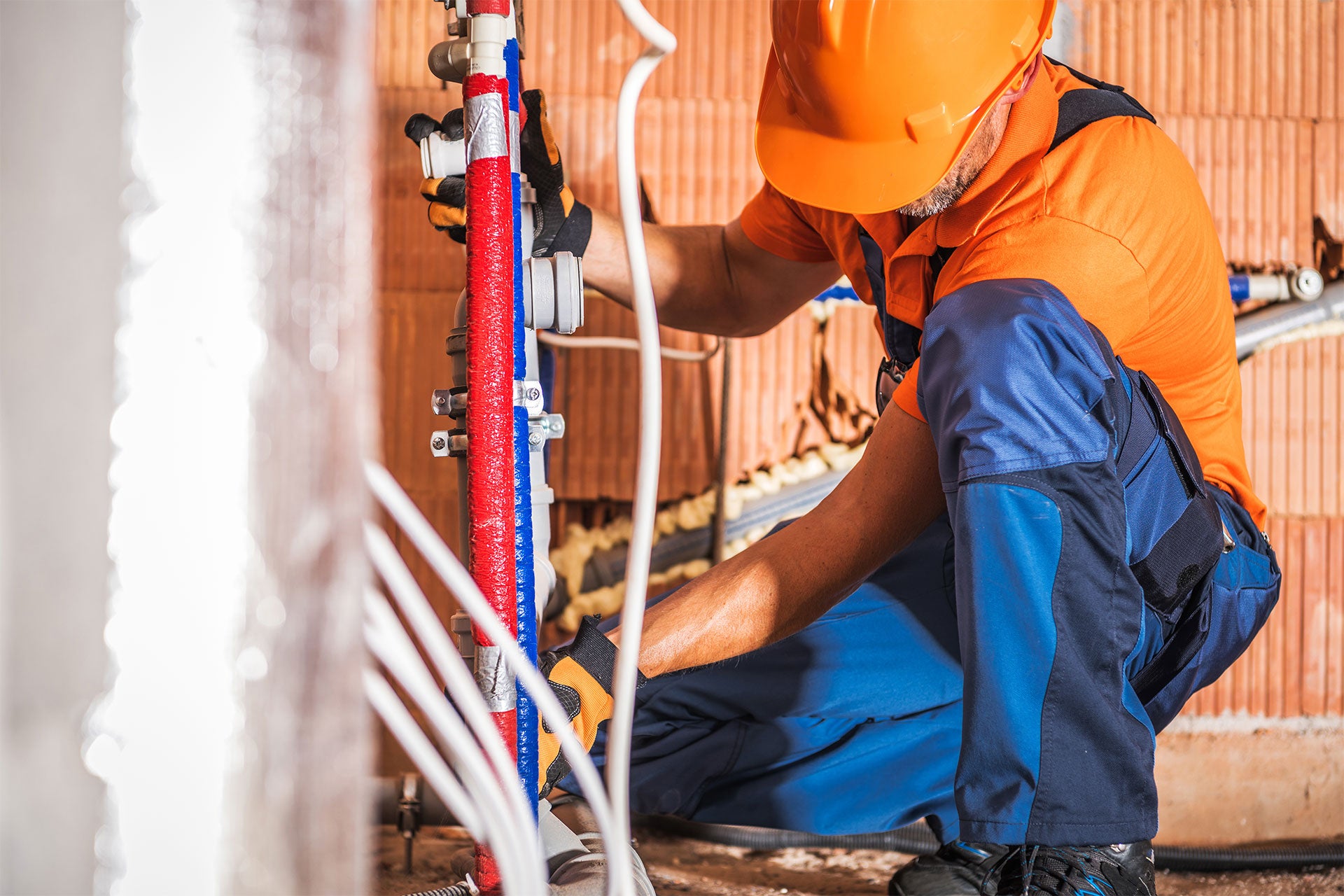 A plumber in an orange hard hat and shirt, and blue pants, connects red and blue insulated pipes to a wall.
