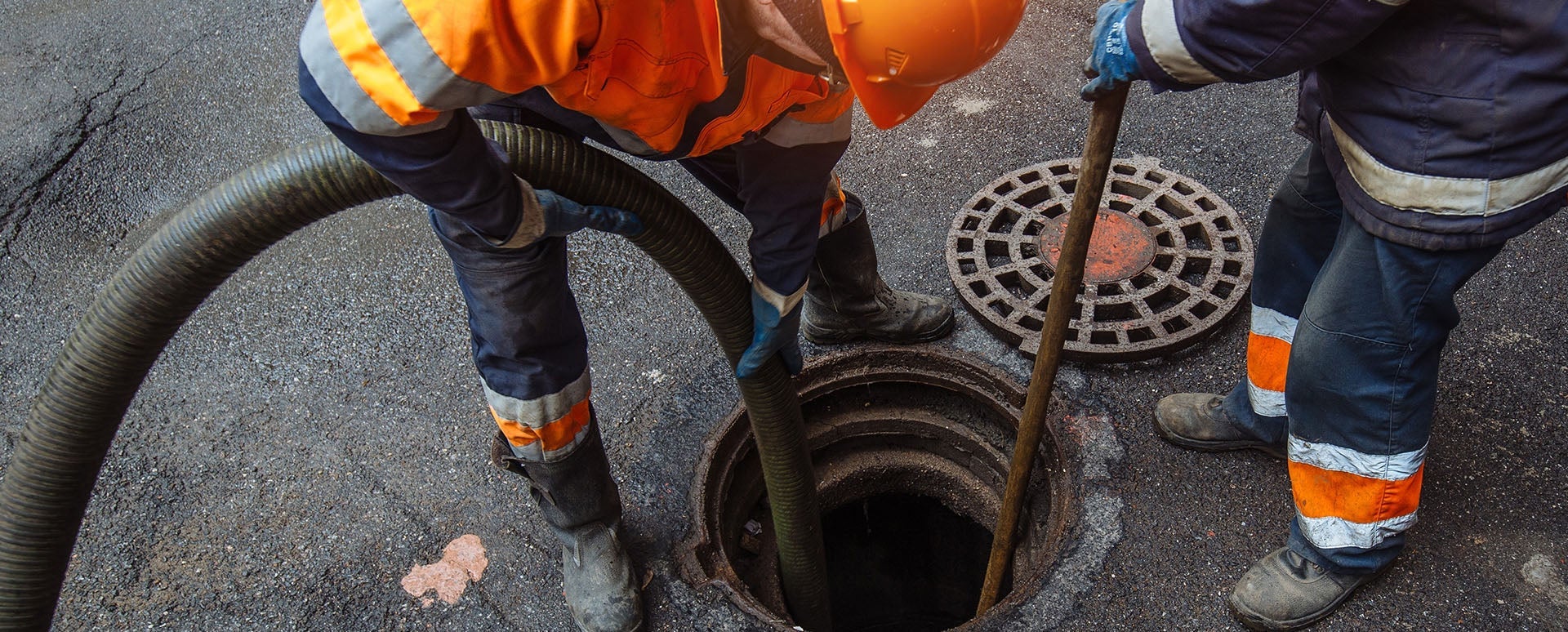 Two workers in safety gear and hard hats clean a sewer, one handling a hose, the other using a pole.