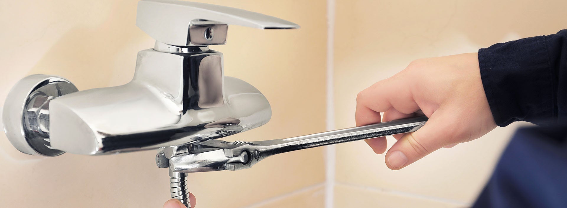 A hand uses a wrench to tighten a chrome faucet on a light-colored tiled wall.