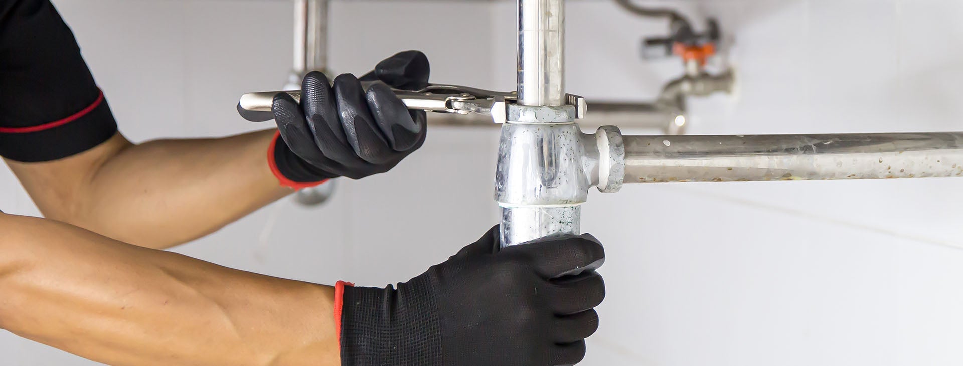 A person in black gloves uses a wrench to tighten a chrome pipe connection under a sink.