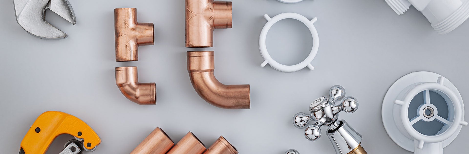Various plumbing tools and pipes, including a wrench, copper fittings, a tap, and white PVC parts on a grey surface.