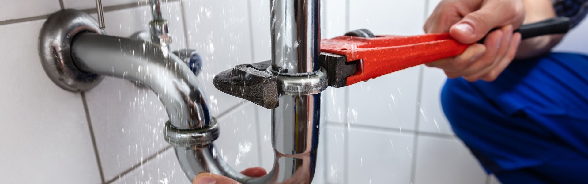 Hands using a red pipe wrench on a leaky chrome pipe, with water splashing.