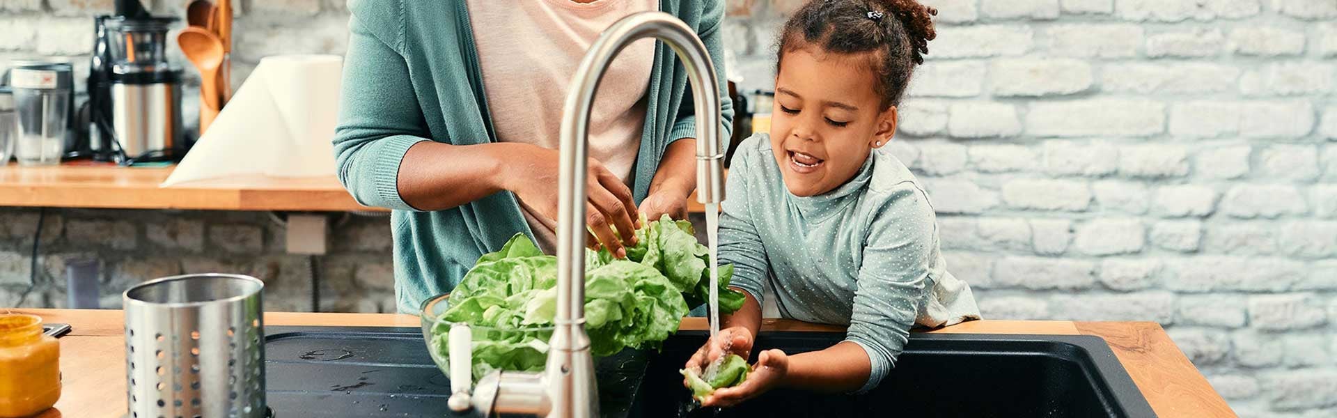 A woman and young child wash lettuce under a kitchen faucet with running water.