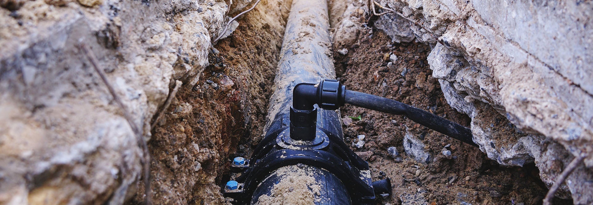 A black pipe, with a connector, runs through a narrow trench of dirt and broken concrete.