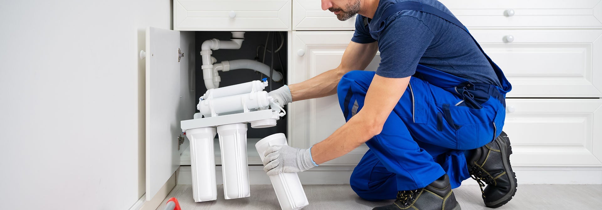 A man in blue overalls and gloves installing a water filter system under a white kitchen sink.