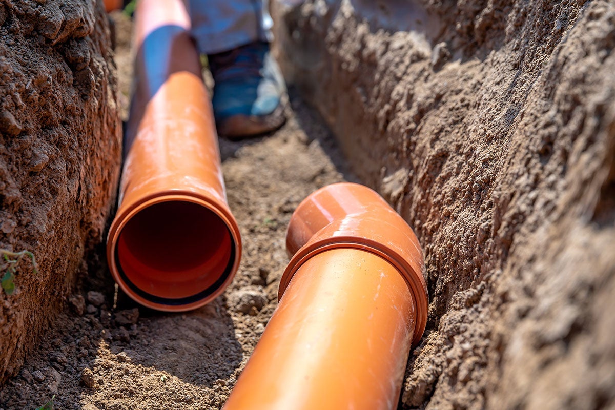Two large orange drainpipes with a bend, lying in a freshly dug dirt trench. A person's blurred feet are visible in the background.