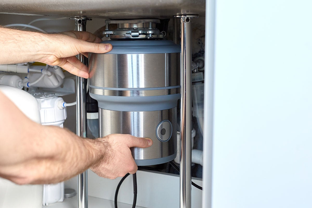 Hands adjusting a stainless steel garbage disposal unit under a kitchen sink.