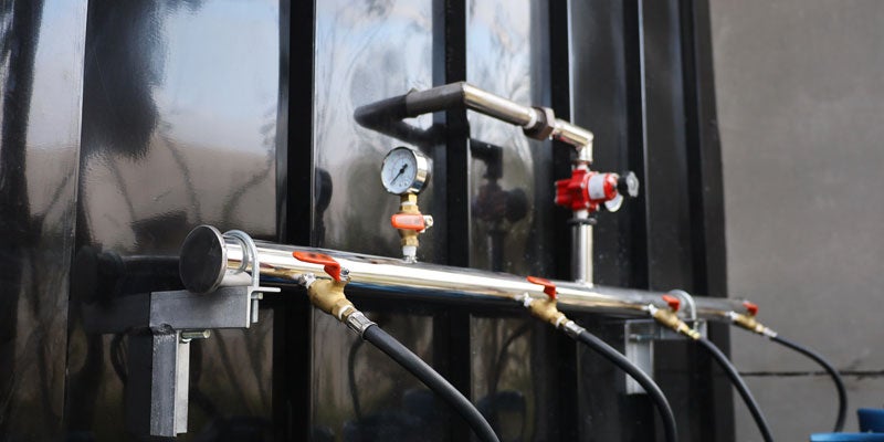 A close-up of shiny chrome plumbing with a pressure gauge, red valves, and black hoses against a dark surface.