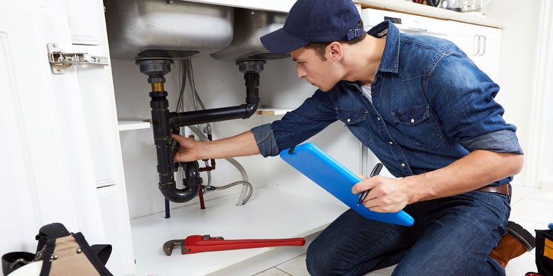 A male plumber in a denim shirt kneels to inspect pipes under a kitchen sink, holding a blue clipboard.