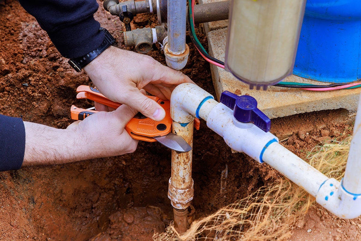A plumber's hands using orange pipe cutters to trim a brown pipe in a muddy trench with various plumbing visible.