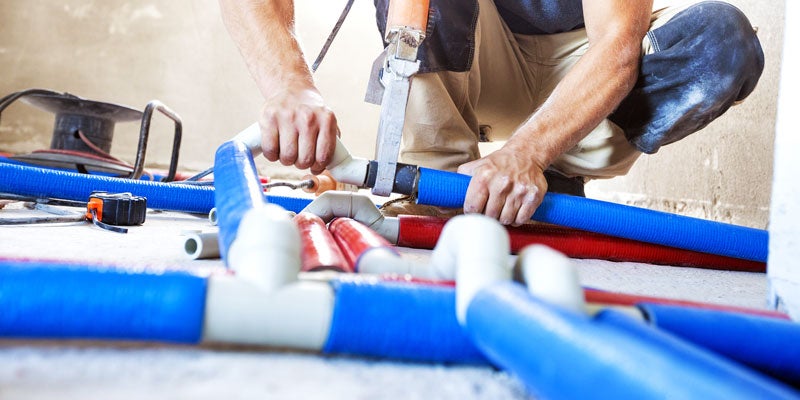 A person kneeling on a floor, installing blue and red PEX pipes, using a heat fusion tool to connect them.