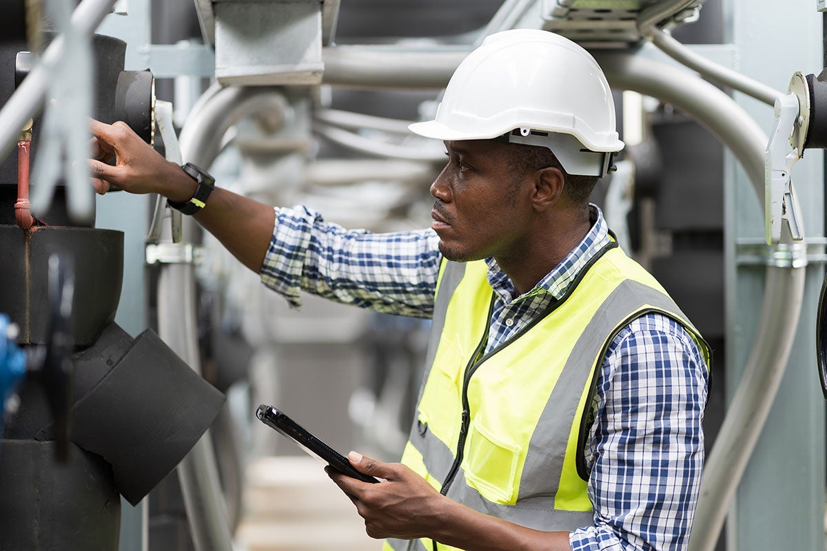 A man in a hard hat and safety vest checks machinery while holding a tablet.