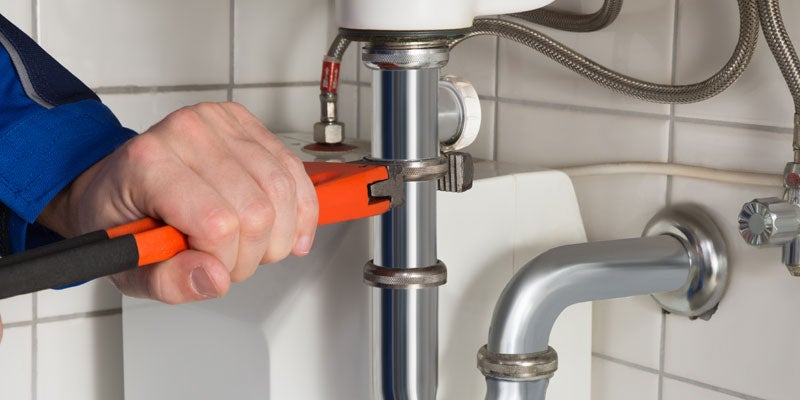 A hand in a blue sleeve uses an orange and black wrench to tighten a chrome pipe under a white sink.