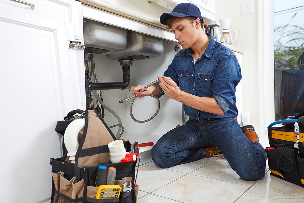 A plumber in a baseball cap and denim outfit kneels and examines a flexible hose under a kitchen sink.
