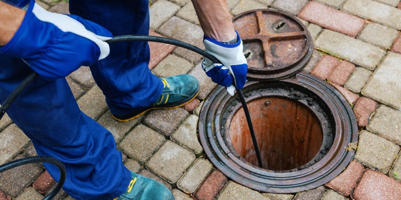 Person in blue work clothes and gloves using plumbing snake to clear a drain or sewer through an open manhole.
