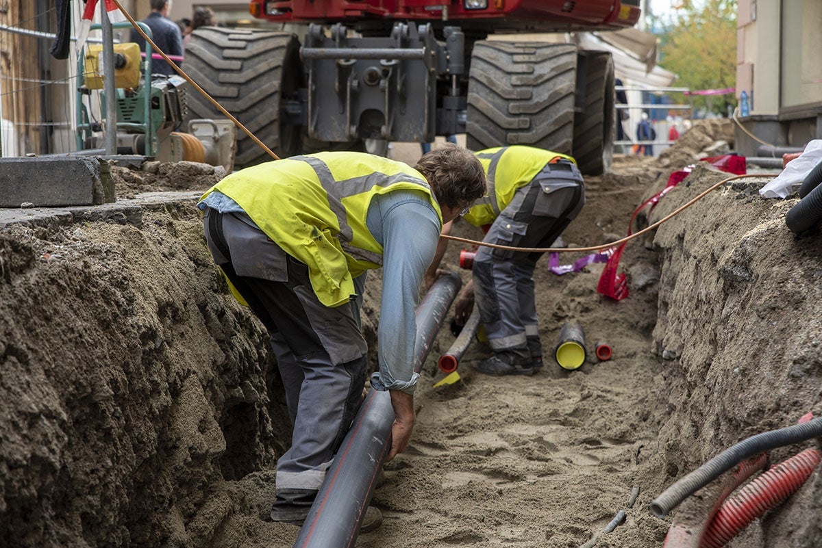 Two construction workers in fluorescent vests lay pipes in a ditch next to a large excavator.
