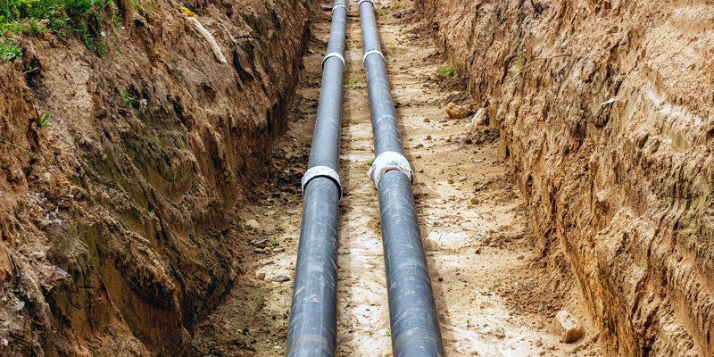 Two dark gray pipes with white connection bands laid in a deep, narrow trench with earthen walls.