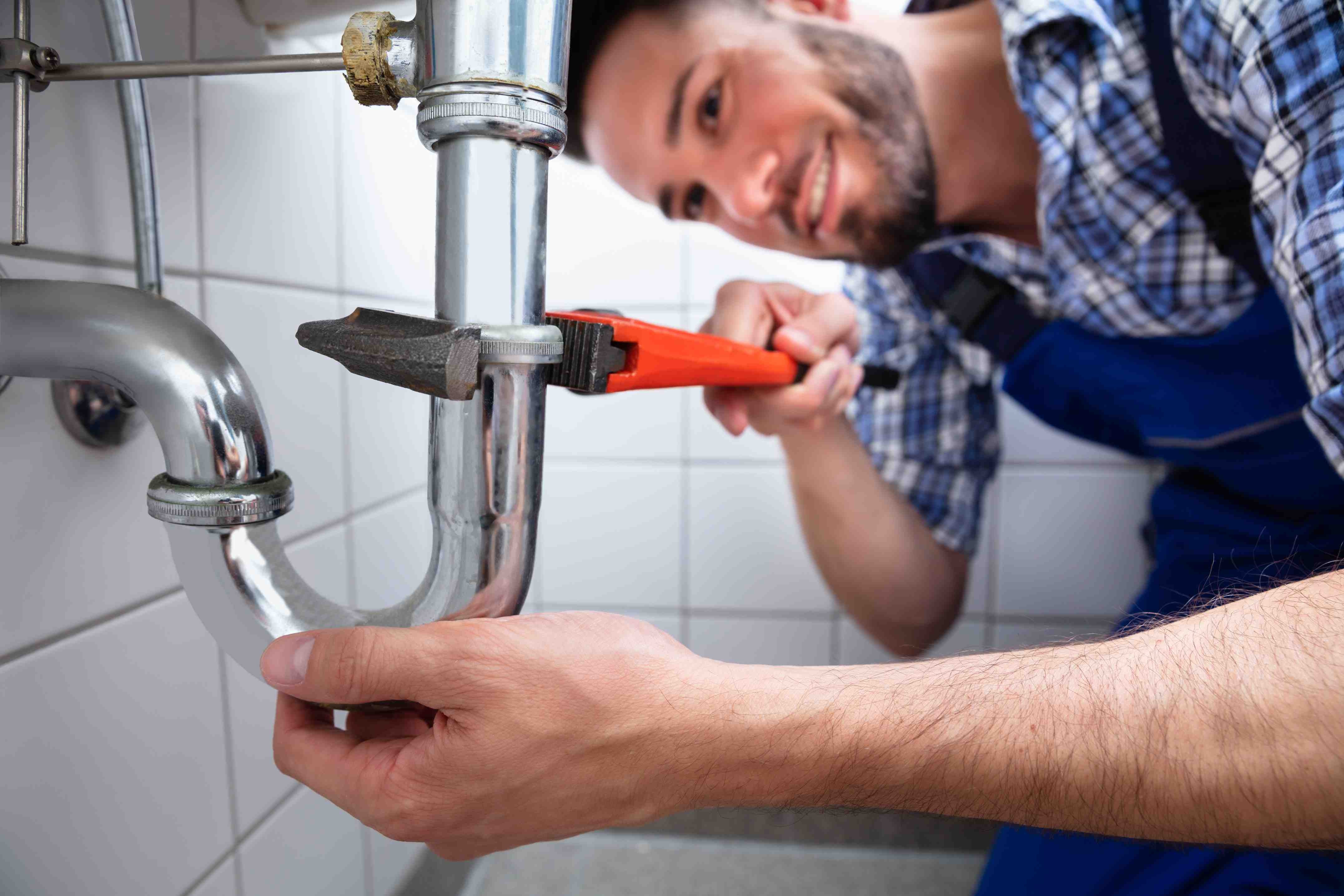 A smiling male plumber in a plaid shirt and blue overalls uses a wrench to tighten a chrome pipe under a sink.